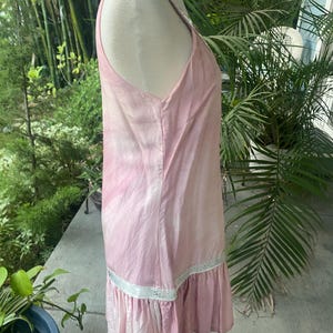 May include: A sleeveless light pink dress featuring a subtle tie-dye pattern, a scoop neckline, and a horizontal white lace trim detail above a gathered ruffle hemline. The garment is displayed on a mannequin, showing a side profile view.