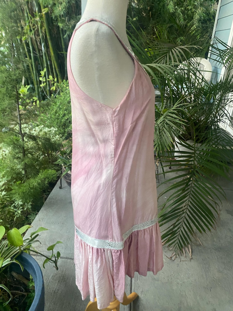 May include: A sleeveless light pink dress featuring a subtle tie-dye pattern, a scoop neckline, and a horizontal white lace trim detail above a gathered ruffle hemline. The garment is displayed on a mannequin, showing a side profile view.