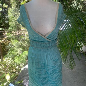 May include: A turquoise dress with a deep V-neckline, short sleeves, and a wide, gathered elasticized waistband. The fabric has a crinkled texture, and the neckline is finished with a light beige trim. The dress falls to a knee-length hem.