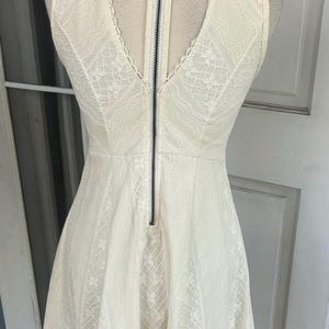 Pu&ograve; includere: Abito in pizzo avorio con scollo a V e gonna ampia. L'abito presenta una cerniera nera sul retro ed &egrave; esposto su un manichino. L'abito &egrave; senza maniche e presenta un delicato motivo in pizzo.