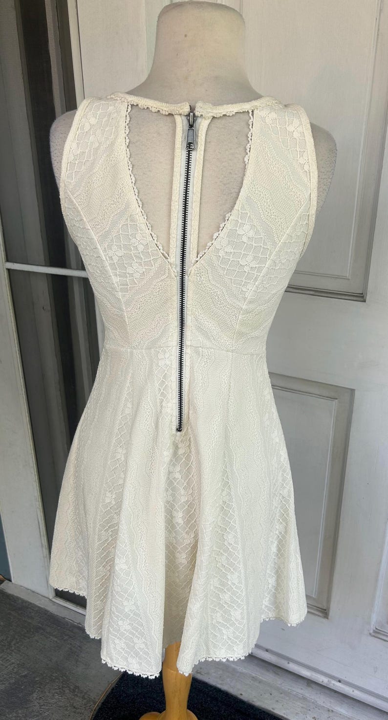Pu&ograve; includere: Abito in pizzo avorio con scollo a V e gonna ampia. L'abito presenta una cerniera nera sul retro ed &egrave; esposto su un manichino. L'abito &egrave; senza maniche e presenta un delicato motivo in pizzo.