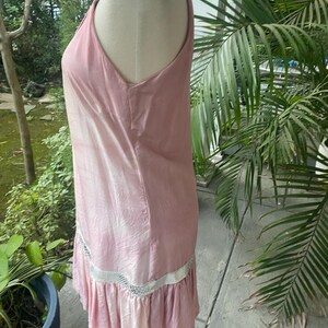 May include: A sleeveless, light pink dress with a subtle sheen and a textured, crinkled appearance. The garment features a horizontal band of white, open-weave mesh trim above a gathered, ruffled hemline that falls to the knee.