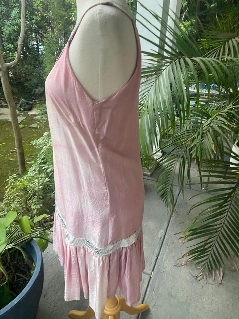 May include: A sleeveless, light pink dress with a subtle sheen and a textured, crinkled appearance. The garment features a horizontal band of white, open-weave mesh trim above a gathered, ruffled hemline that falls to the knee.
