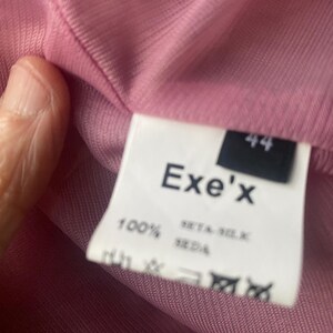May include: A pink ribbed garment featuring a white interior clothing label. The label displays the brand name Exe'x, the size 44, and the text 100% SETA-SILK SEDA, indicating the material composition, along with several standard garment care symbols.