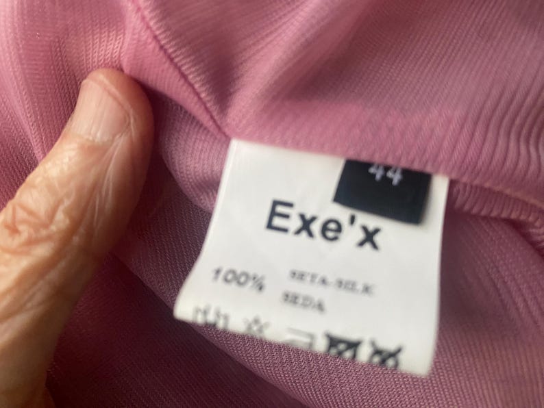 May include: A pink ribbed garment featuring a white interior clothing label. The label displays the brand name Exe'x, the size 44, and the text 100% SETA-SILK SEDA, indicating the material composition, along with several standard garment care symbols.