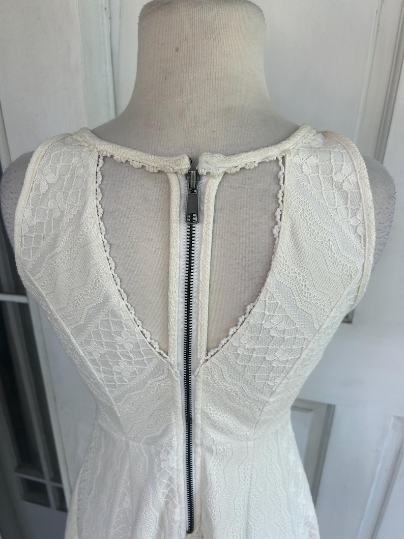 Pu&ograve; includere: Abito senza maniche color avorio con sovrapposizione in pizzo. L'abito presenta una schiena a V con un design a chiave e una cerniera nera a tutta lunghezza. L'abito &egrave; esposto su un manichino.