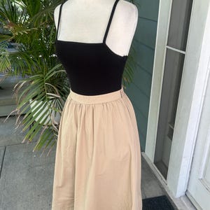 May include: A black ribbed tank top with thin straps is paired with a tan, knee-length skirt. The skirt has a gathered waist and a flowing silhouette. The outfit is displayed on a mannequin, showcasing a casual, everyday style.