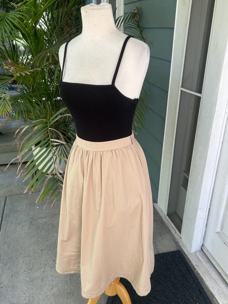 May include: A black ribbed tank top with thin straps is paired with a tan, knee-length skirt. The skirt has a gathered waist and a flowing silhouette. The outfit is displayed on a mannequin, showcasing a casual, everyday style.