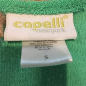 May include: Close-up of a green garment tag with the brand name "capelli new york" in yellow. The tag also includes care instructions and the size "S". The garment is a light green color.