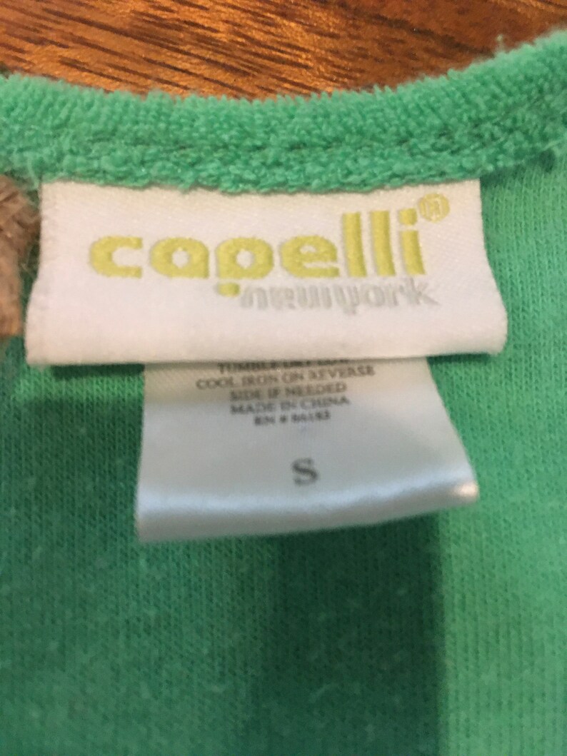 May include: Close-up of a green garment tag with the brand name "capelli new york" in yellow. The tag also includes care instructions and the size "S". The garment is a light green color.