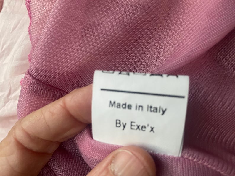 May include: A pink sheer mesh garment featuring a white rectangular fabric label. The label displays black text that reads Made in Italy and By Exe'x, positioned above a series of small black laundry care symbols printed on the white tag.