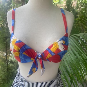 May include: A bikini top featuring a vibrant floral pattern with red, blue, and yellow colors. The garment has thin shoulder straps, a sweetheart neckline, and a central tie detail at the front, displayed on a mannequin torso.