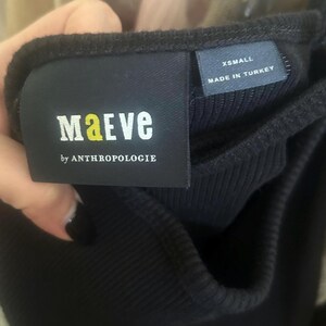 May include: Close-up of a black ribbed tank top with a tag that reads "MAeVE by ANTHROPOLOGIE". The tag also indicates the size is "XSMALL" and "MADE IN TURKEY". The tank top has adjustable straps.
