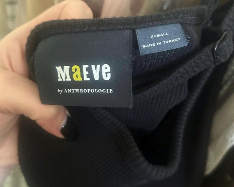 May include: Close-up of a black ribbed tank top with a tag that reads "MAeVE by ANTHROPOLOGIE". The tag also indicates the size is "XSMALL" and "MADE IN TURKEY". The tank top has adjustable straps.