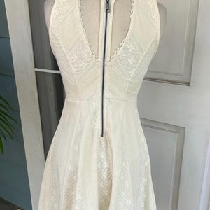 Pu&ograve; includere: Abito in pizzo avorio con scollo a V e cerniera nera sulla schiena. L'abito &egrave; senza maniche e ha una gonna svasata. L'abito &egrave; esposto su un manichino.
