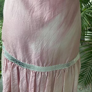May include: A light pink dress with a crinkled texture and a white, diagonal tie-dye pattern. The garment features a horizontal band of white, textured snakeskin-patterned trim above a pleated ruffle hemline that extends around the lower section.