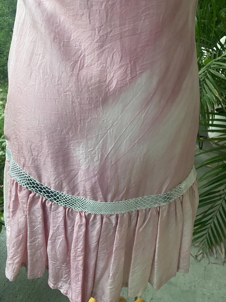 May include: A light pink dress with a crinkled texture and a white, diagonal tie-dye pattern. The garment features a horizontal band of white, textured snakeskin-patterned trim above a pleated ruffle hemline that extends around the lower section.