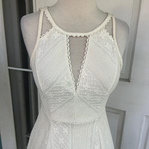 Pu&ograve; includere: Un abito di pizzo bianco con scollatura all'americana e scollo a V. L'abito presenta un corpetto aderente e una gonna svasata, che mostra intricati motivi in pizzo. L'abito &egrave; esposto su un manichino.