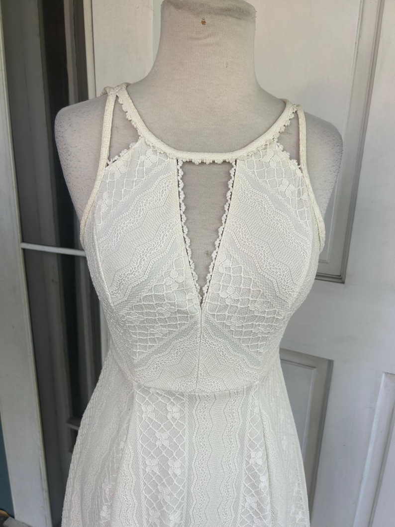 Pu&ograve; includere: Un abito di pizzo bianco con scollatura all'americana e scollo a V. L'abito presenta un corpetto aderente e una gonna svasata, che mostra intricati motivi in pizzo. L'abito &egrave; esposto su un manichino.