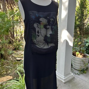 May include: Black sleeveless dress with a unique art print on the front. The print features a portrait with green, blue, and peach tones. The dress has a simple, elegant design, perfect for a casual or semi-formal occasion.