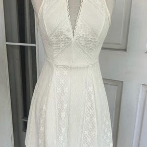 Pu&ograve; includere: Un abito di pizzo bianco con scollatura all'americana e scollo a V. L'abito presenta un corpetto aderente e una gonna svasata. Gli intricati dettagli in pizzo ricoprono l'intero abito. L'abito &egrave; esposto su un manichino.