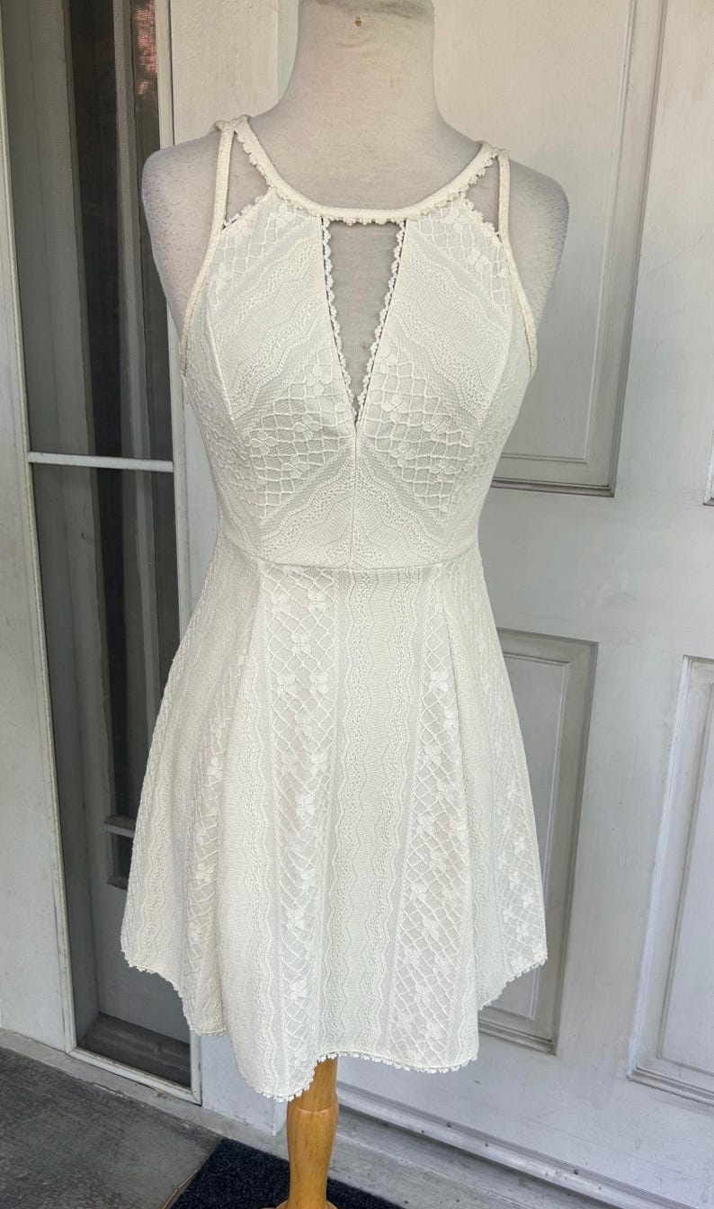 Pu&ograve; includere: Un abito di pizzo bianco con scollatura all'americana e scollo a V. L'abito presenta un corpetto aderente e una gonna svasata. Gli intricati dettagli in pizzo ricoprono l'intero abito. L'abito &egrave; esposto su un manichino.
