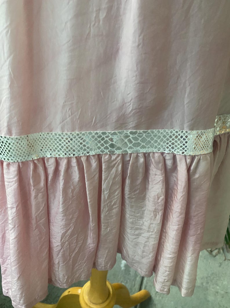 May include: A light pink garment with a crinkled texture featuring a horizontal band of white snakeskin-patterned trim above a gathered ruffle hem. The fabric has a subtle sheen and the design includes a distinct horizontal seam detail.