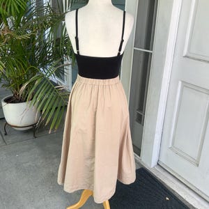 May include: A black camisole top paired with a beige midi skirt, displayed on a mannequin. The skirt has a gathered waist and a flared silhouette. The outfit is presented on a wooden stand, with a green plant and a white door in the background.