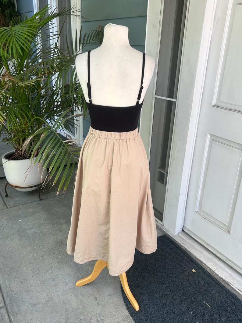 May include: A black camisole top paired with a beige midi skirt, displayed on a mannequin. The skirt has a gathered waist and a flared silhouette. The outfit is presented on a wooden stand, with a green plant and a white door in the background.