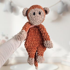 May include: A crocheted plush monkey toy with brown fur and a beige face. The monkey has a long tail and is holding its arms out.