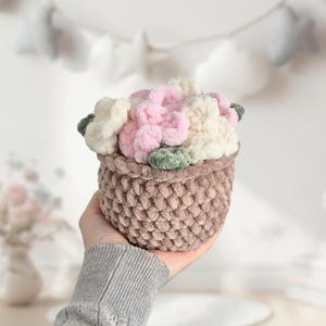 May include: A small, brown, crocheted basket filled with plush, light pink and white flowers, and green leaves. The basket is held in a hand, showcasing the handmade floral arrangement. The overall theme is a decorative, handcrafted item.
