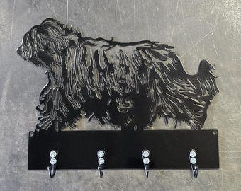 Pode incluir: Porta-chaves de metal preto com uma silhueta de cão estilizada. O porta-chaves tem quatro ganchos abaixo do desenho do cão. O pelo do cão é detalhado com linhas recortadas. O porta-chaves é montado numa superfície metálica.