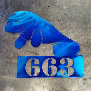 May include: A blue metal manatee sculpture with the number 663 cut out below. The manatee is a vibrant blue with a brushed metal finish. The number plate has two holes for mounting.