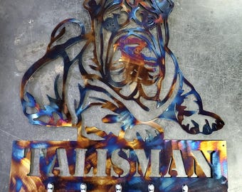 Pode incluir: Arte de parede em metal com um design de bulldog estilizado com um porta-chaves. A obra de arte é em tons de azul, dourado e bronze. A palavra "TALISMAN" é recortada abaixo do cão, com cinco ganchos para chaves.