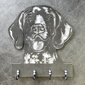 May include: A metal key holder featuring a dog's head silhouette with a happy expression. The key holder has four black hooks below the dog's face. The metal is a dark gray color.