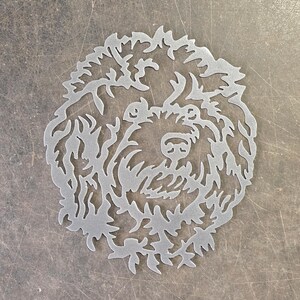 Puede incluir: Un recorte de metal plateado de la cara de un perro, con detalles intrincados de pelaje y rasgos faciales. El diseño es una representación estilizada de un perro, probablemente un caniche o raza similar, adecuado para decoración de pared.