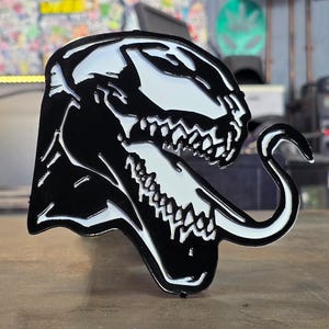 May include: Black and white enamel pin featuring a stylized depiction of a monster's head with a long, curved tongue. The design has a glossy finish and is set against a blurred background.