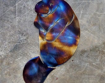 May include: A metal manatee sculpture with a vibrant, heat-treated finish. The colors range from deep blues and purples to warm oranges and browns. The manatee is depicted in a side profile, with its flipper resting on its chest.