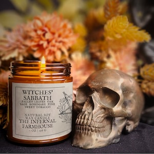 May include: A brown glass candle jar with a white label that reads "Witches' Sabbath" and lists the scent notes: "Fallen Leaves Oak, Sage Rosemary Pine, Sweet Tobacco". The label also states "Natural Soy Wax Candle, The Infernal Farmhouse, 7 oz | 198 g". The candle is sitting on a dark surface next to a white skull.