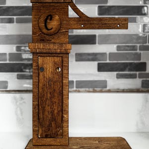 Puede incluir: Un llavero de madera marrón oscuro. El diseño incluye una parte superior decorativa, un círculo con la letra "C" y una sección inferior en forma de puerta. Un brazo horizontal con dos agujeros para colgar llaves.