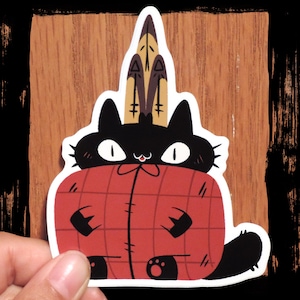 May include: A sticker featuring a black cat with white eyes and a red blanket. The cat wears a headdress of feathers. The blanket has a plaid pattern. The sticker is held by a hand against a wooden background.
