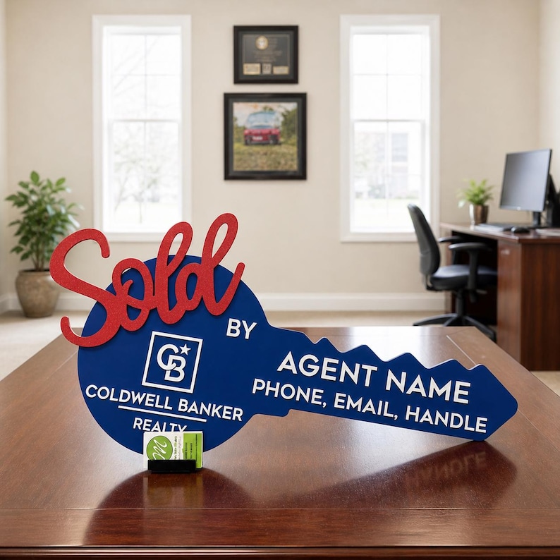 May include: A blue key-shaped sign with the red script word Sold, the Coldwell Banker Realty logo, and white text reading BY AGENT NAME, PHONE, EMAIL, HANDLE. A small business card holder is attached to the bottom of the blue key-shaped sign.