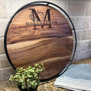 May include: A round wooden serving board with a black metal frame. The board features the engraved text "MORAN FAMILY" with a decorative initial "M". A small potted plant and a blue and white striped towel are in the foreground.