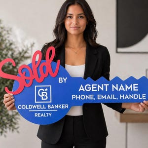 May include: A large blue key-shaped sign with the words "Sold" in red script. The sign also features the Coldwell Banker Realty logo and space for an agent's name, phone, email, and handle. The sign is held by a person.