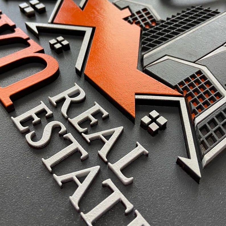 May include: A three-dimensional wall sign featuring the text REAL ESTATE in white raised lettering. An orange roof-shaped icon sits above the text, accompanied by a dark gray grid-patterned building graphic and small square accents on a dark gray base.