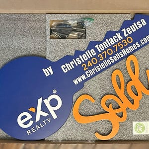 May include: A large blue key-shaped sign with the white exp REALTY logo and the text Christelle Tonfack Zeutsa 240.370.7530 www.ChristelleSellsHomes.com, accompanied by an orange cursive Sold sign, all packed in a box with grey foam inserts.