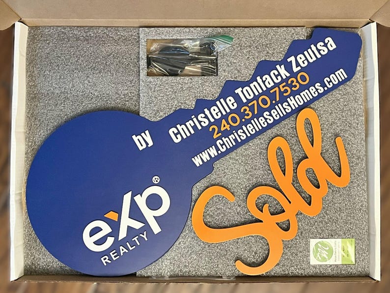 May include: A large blue key-shaped sign with the white exp REALTY logo and the text Christelle Tonfack Zeutsa 240.370.7530 www.ChristelleSellsHomes.com, accompanied by an orange cursive Sold sign, all packed in a box with grey foam inserts.