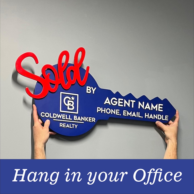 May include: A large blue key-shaped sign featuring the red script word Sold, the Coldwell Banker Realty logo, and white text reading BY AGENT NAME, PHONE, EMAIL, HANDLE. The sign is designed for display in a real estate office setting.