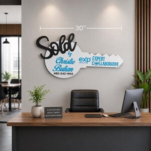 May include: A 30-inch wide white key-shaped wall sign with black script text reading Sold by, blue text reading Christie Bulian, 480-242-1943, exp REALTY, and EXPERT COLLABORATIVE. The sign is mounted on a wall above a desk and office chair.