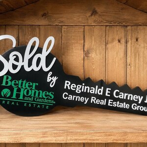 May include: A large black key-shaped sign featuring the white text Sold by, the green Better Homes and Gardens Real Estate logo, and the white text Reginald E Carney Jr Carney Real Estate Group printed across the blade of the key.